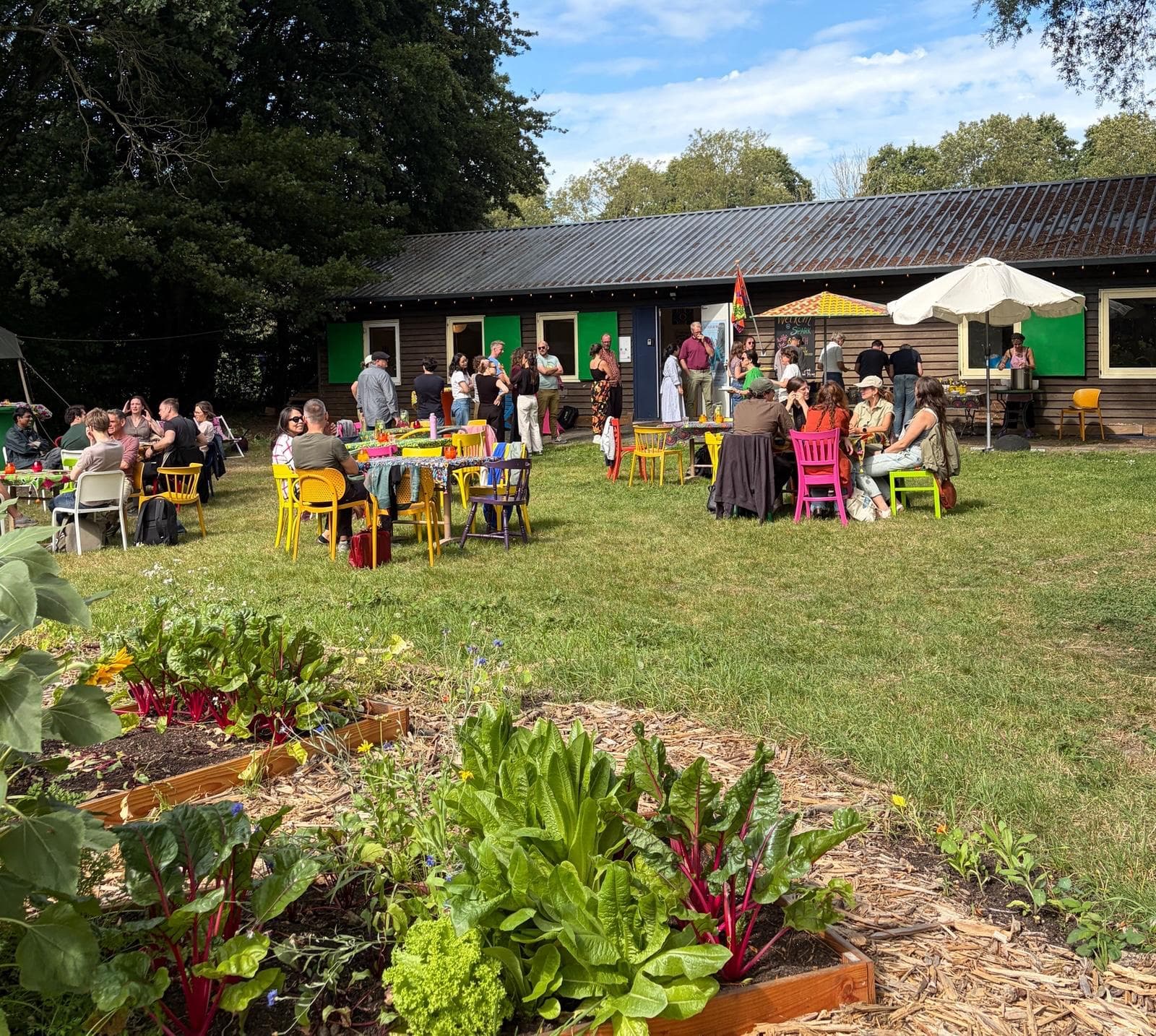 Mensen aan kleurrijke tafels op het gras bij een houten gebouw met moestuinbakken op de voorgrond