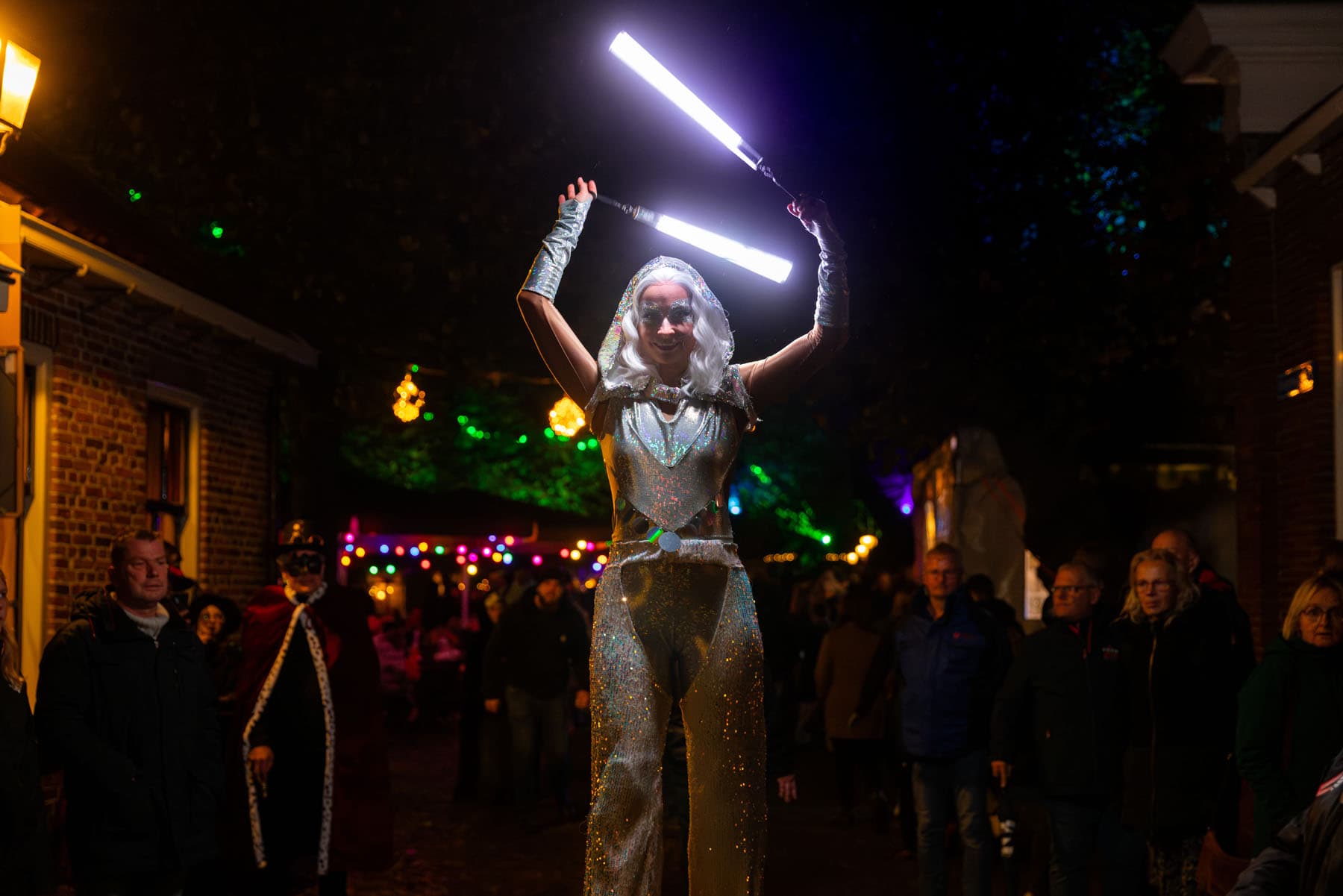 Steltloper in glinsterend zilveren kostuum met lichtstaven in een avondlijk verlichte straat van de vesting